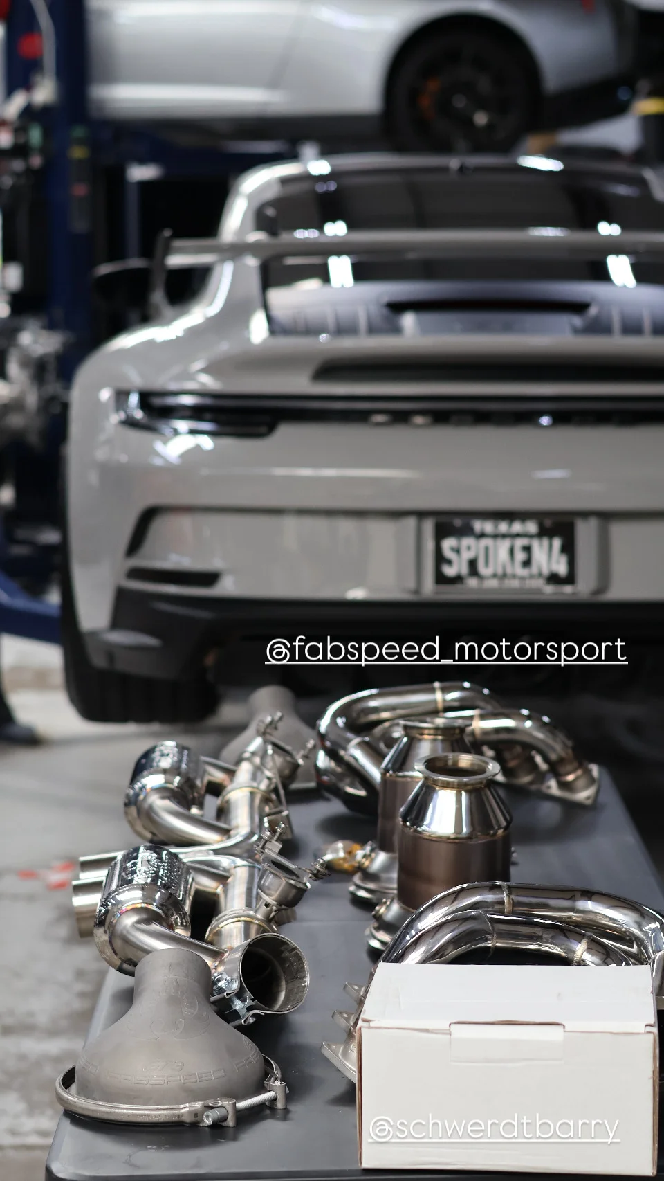 Fabspeed Motorsport exhaust system components laid out on a table in a performance shop with a Porsche on a lift in the background.