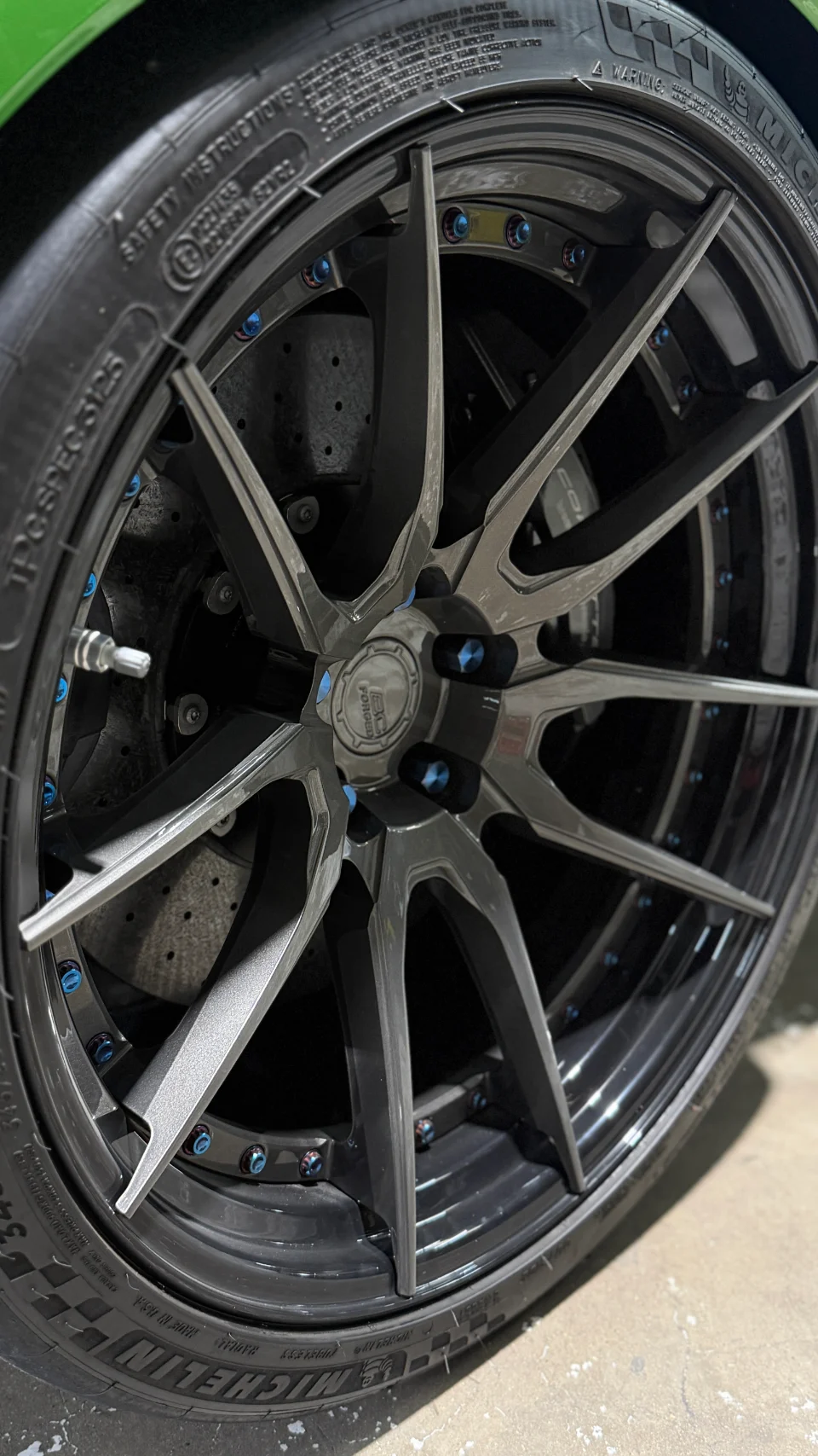 Close-up of a custom multi-spoke forged wheel with blue lug nuts, performance tire, and large brake caliper on a green car.