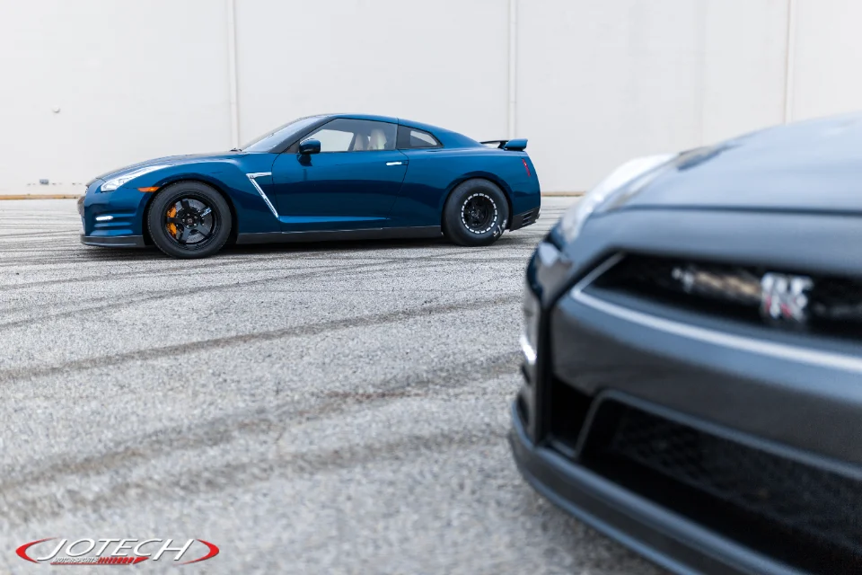 A blue Nissan GT-R with drag wheels and orange brake calipers parked on asphalt with tire marks, with another GT-R in the foreground.