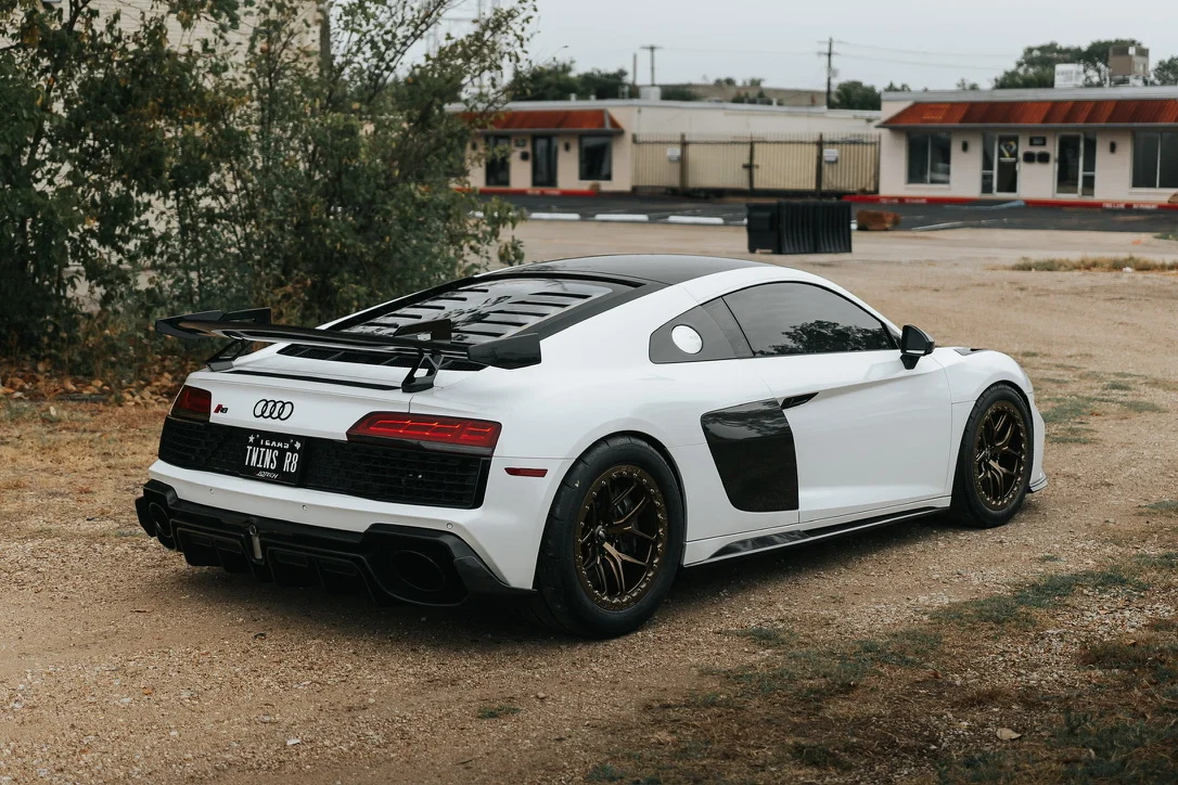 A white Audi R8 with aftermarket bronze wheels and a large carbon fiber rear wing parked on a gravel lot.