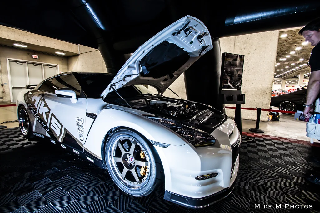 A white Nissan GT-R with an HKS livery and its hood open, displaying the engine bay at an indoor car show.