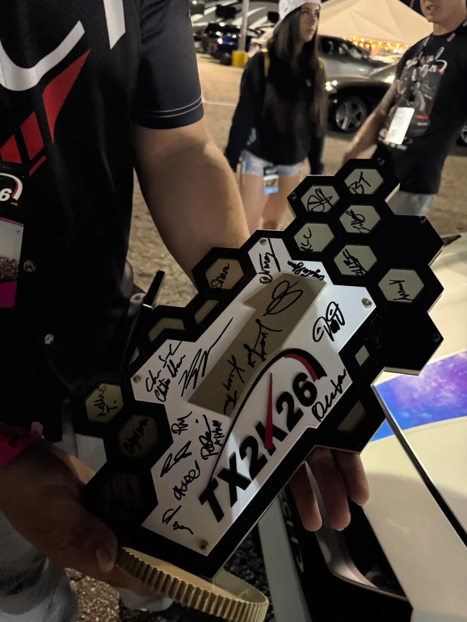 Person holding a signed TX2K26 event trophy at an outdoor car event, with other attendees in the background.