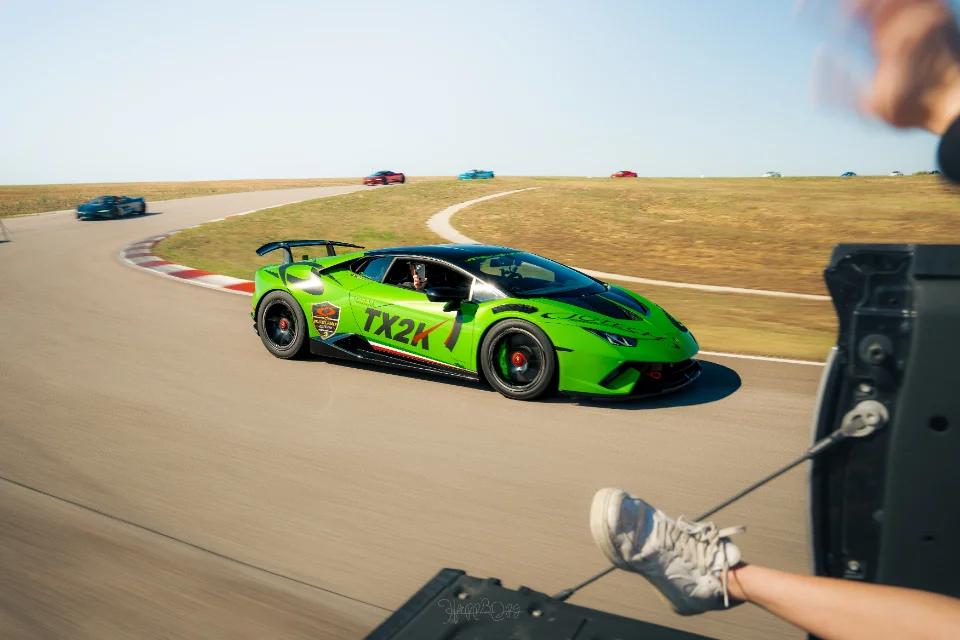 A vibrant green Lamborghini Huracan with TX2K livery speeds around a race track, captured from a moving vehicle.