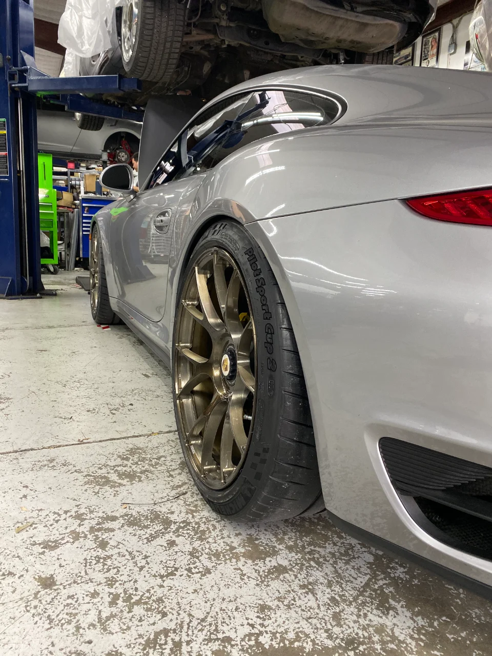 A silver Porsche 911 on a lift in a shop, showcasing aftermarket bronze wheels and Michelin Pilot Sport Cup 2 tires.