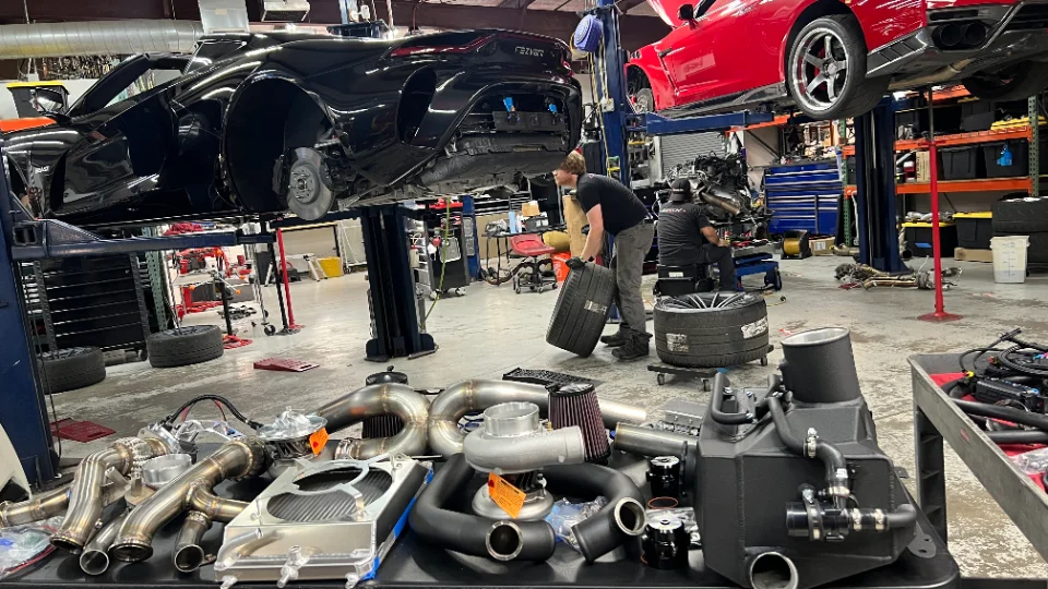 Mechanics working on a black McLaren and a red Camaro on lifts in a performance tuning shop, with a table full of turbocharger kits and other parts in the foreg