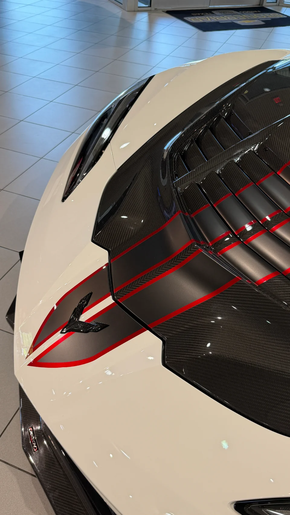 Close-up of a white Corvette C8 rear deck with carbon fiber accents, red and black racing stripes, and a Jotech logo, in a showroom.