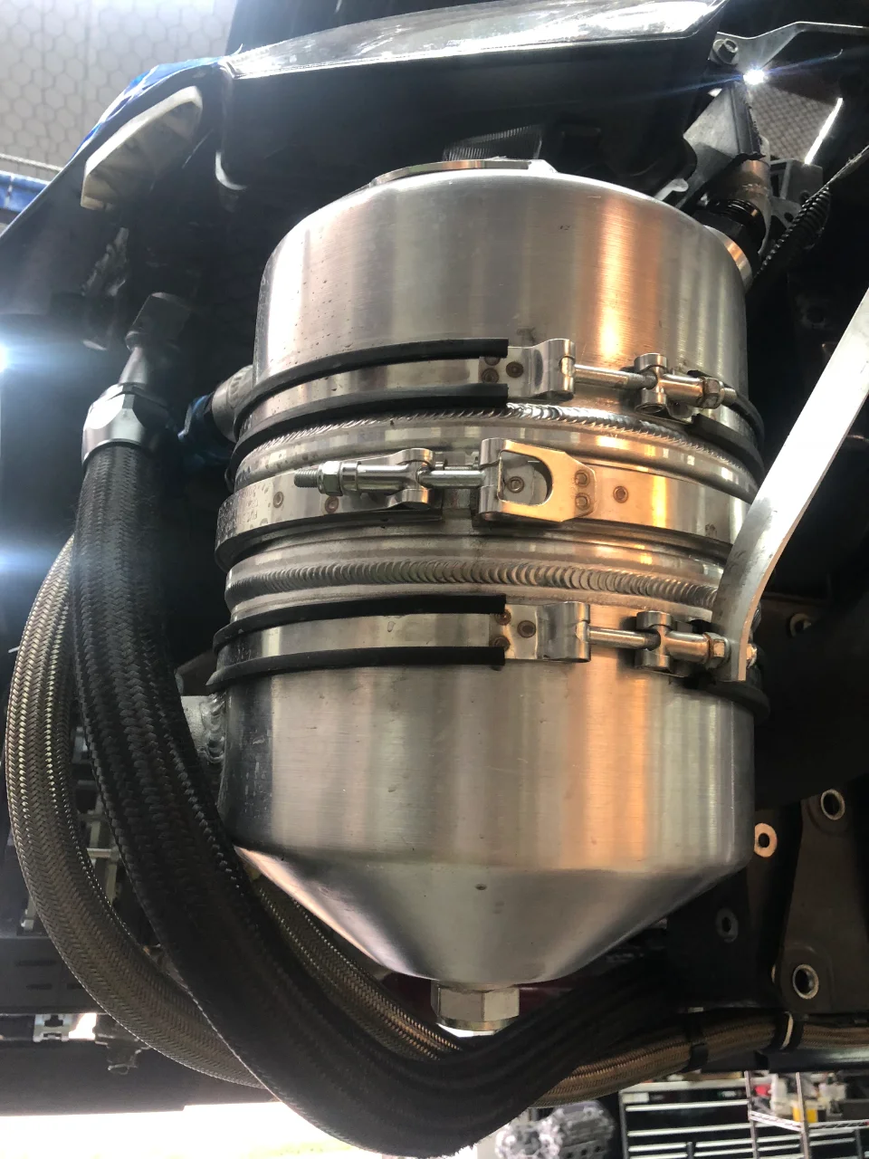 Close-up of an aftermarket aluminum fuel surge tank with braided lines and clamps installed in a performance car's engine bay.