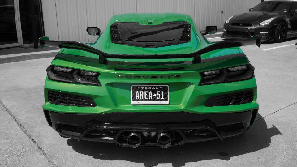 A vibrant green Chevrolet Corvette C8 with a large aftermarket rear wing and custom 'AREA 51' Texas license plate, parked with a black Nissan GT-R in the backgr