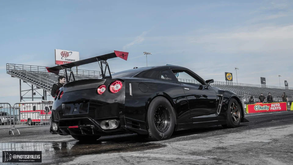 A black Nissan GT-R with a large rear wing and drag tires at a drag strip, viewed from the rear.