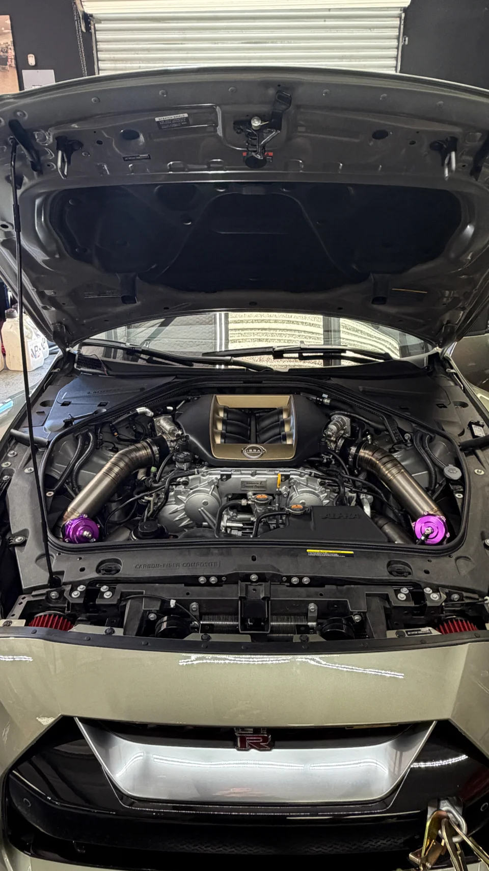 A detailed shot of a Nissan GT-R R35 engine bay showcasing aftermarket turbo piping, purple blow-off valves, and an upgraded intake manifold.