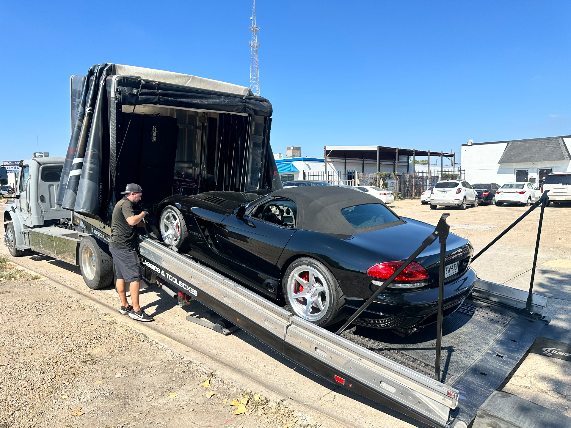 A black Dodge Viper convertible being unloaded from a specialized enclosed car hauler by a technician under a clear sky.