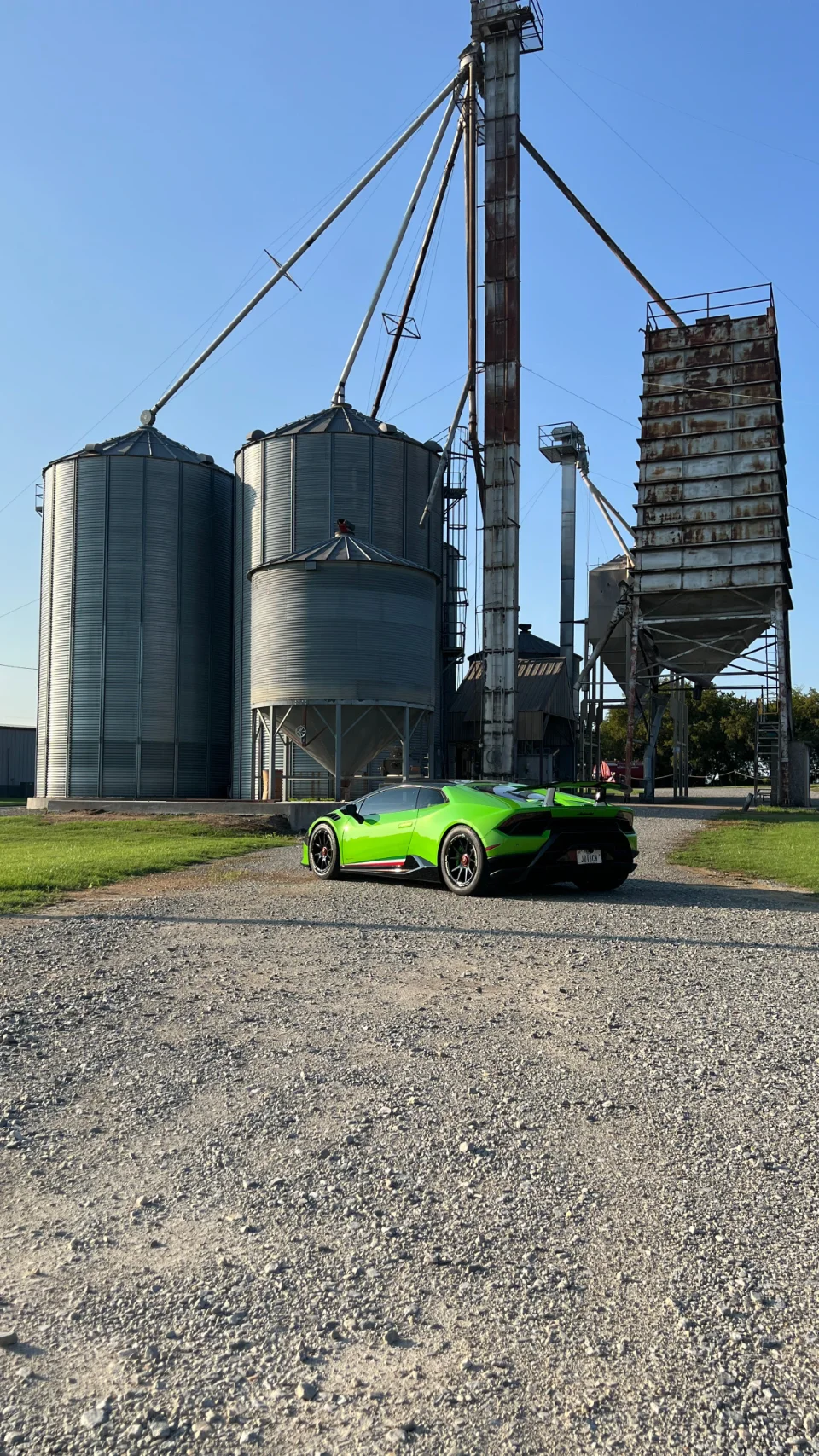 A vibrant green Lamborghini supercar parked on a gravel road next to large grain silos under a clear blue sky.