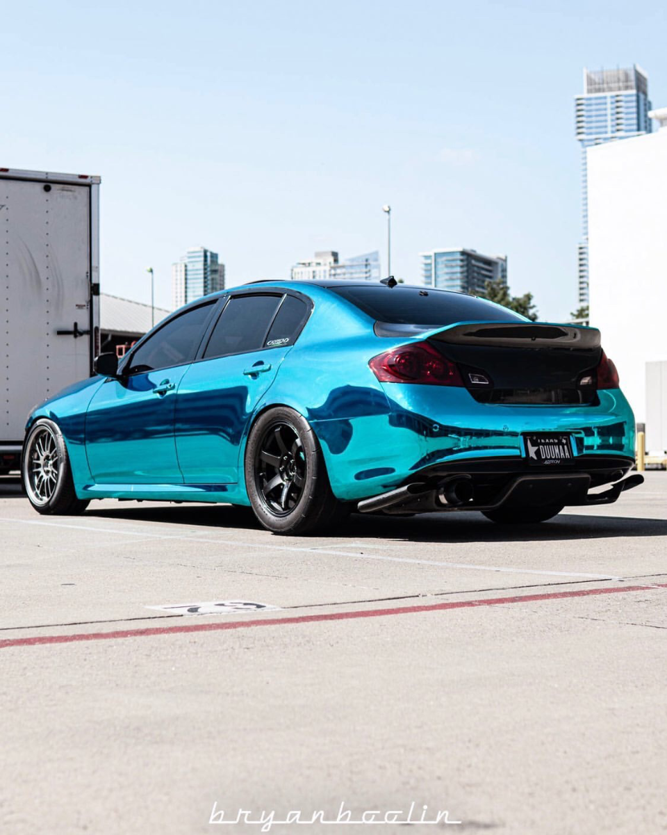 A teal chrome wrapped Infiniti G37 sedan with aftermarket wheels, rear spoiler, and custom exhaust system parked outdoors with a city skyline.