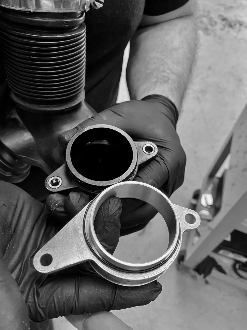 A mechanic's gloved hands hold an OEM black plastic intake pipe next to a shiny aftermarket metal induction adapter.