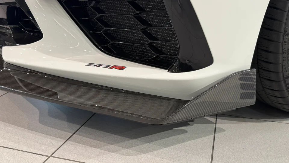 A close-up of a white car's front bumper featuring a carbon fiber splitter and an SBR logo.