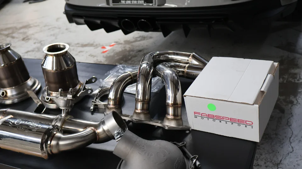 Fabspeed Motorsport exhaust system components including headers, catalytic converters, and mufflers laid out in a shop with a Porsche rear bumper in the backgro