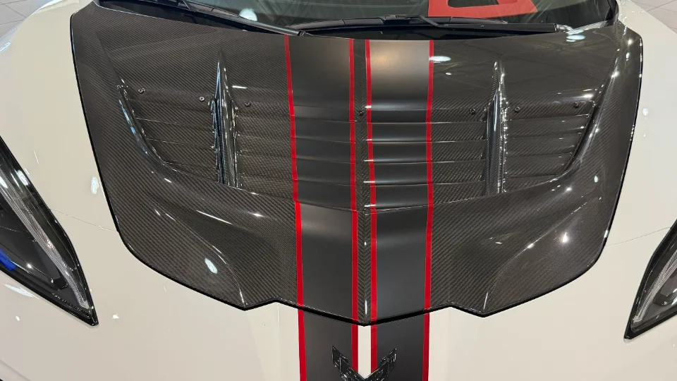 A close-up of a white C8 Corvette's carbon fiber hood featuring black and red racing stripes and functional vents.