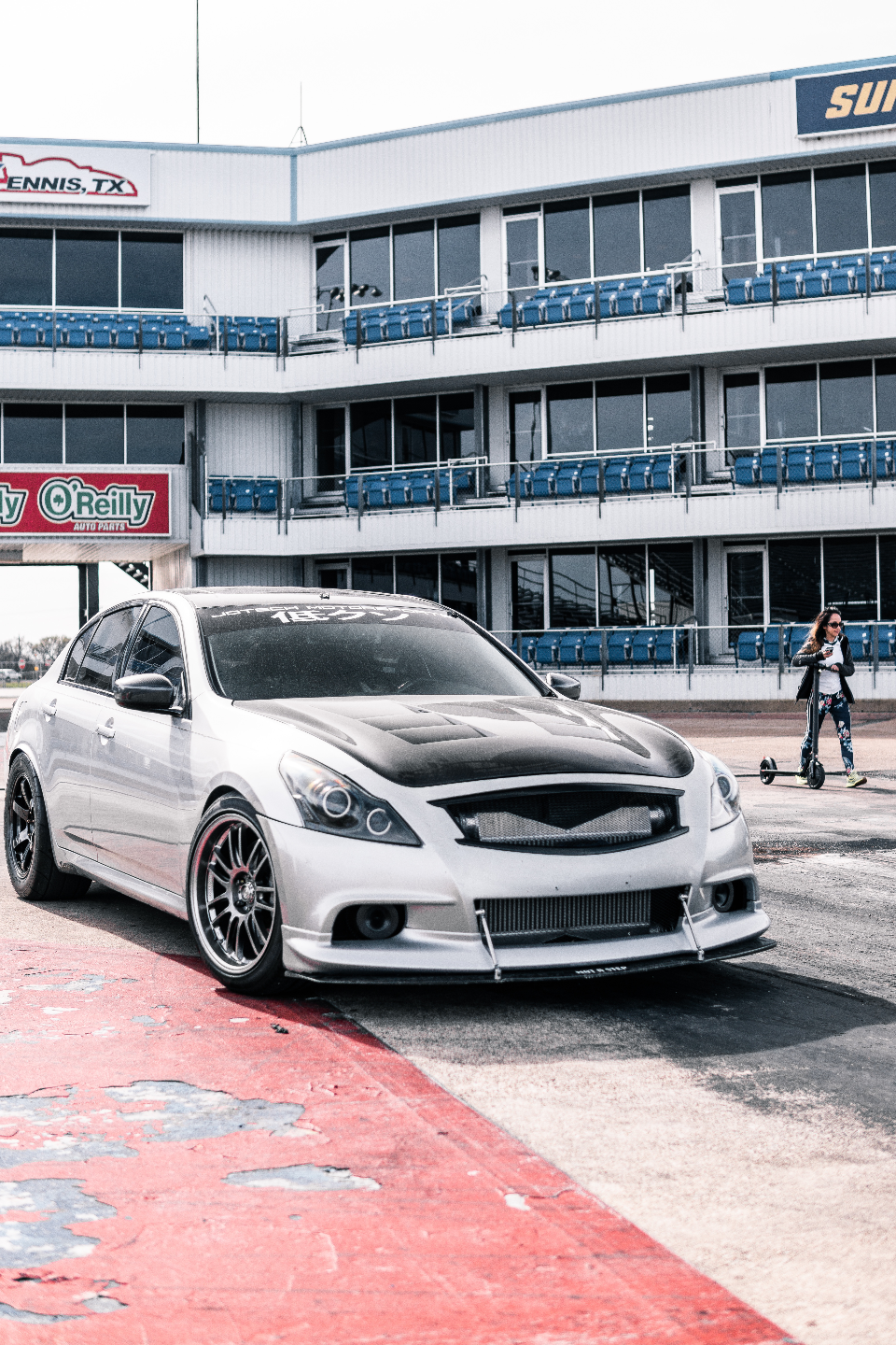 Silver Infiniti G37 sedan with a carbon fiber hood and aftermarket wheels at a drag strip with a grandstand in the background.