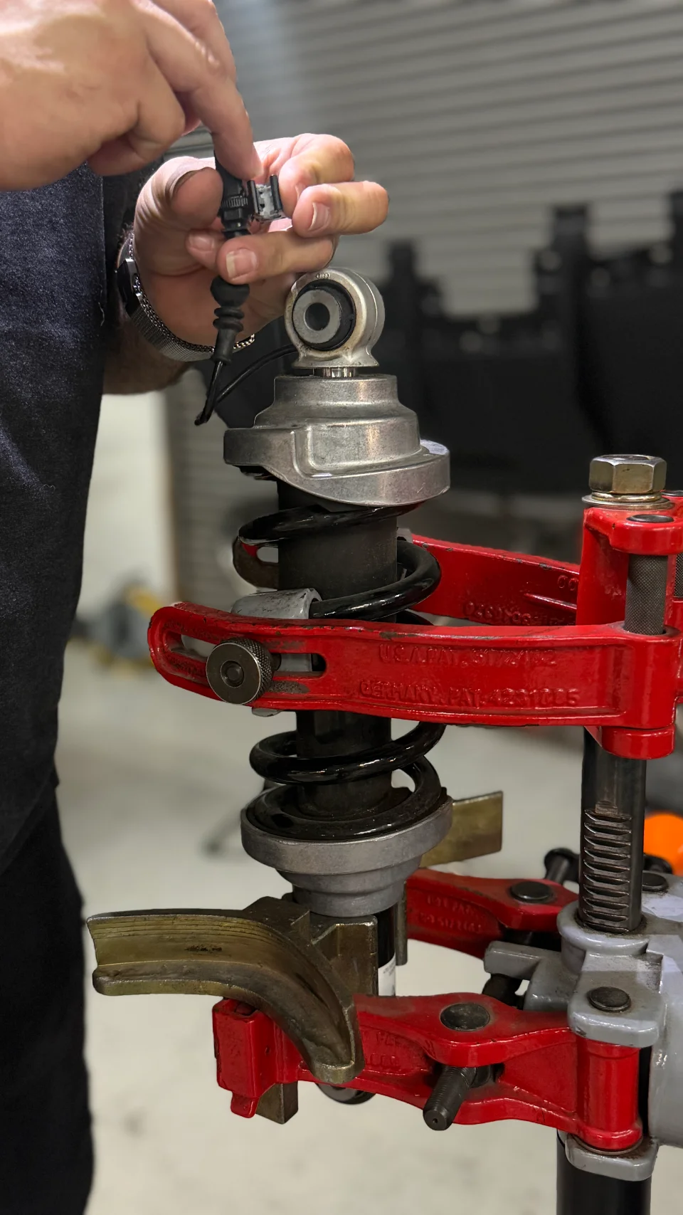A mechanic's hands working on a car's suspension coilover using a spring compressor tool in a shop.
