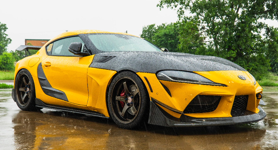 A yellow Toyota Supra with a widebody kit, carbon fiber hood, and aftermarket wheels parked on a wet surface.