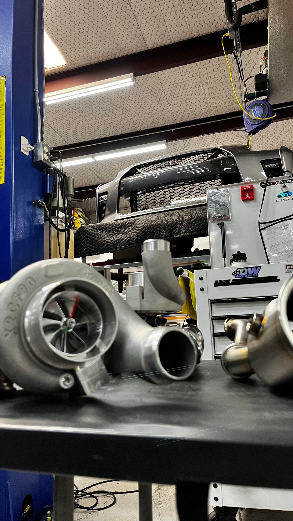 A large turbocharger and associated piping on a workbench in a performance tuning shop, with a Nissan GT-R bumper on a lift in the background.
