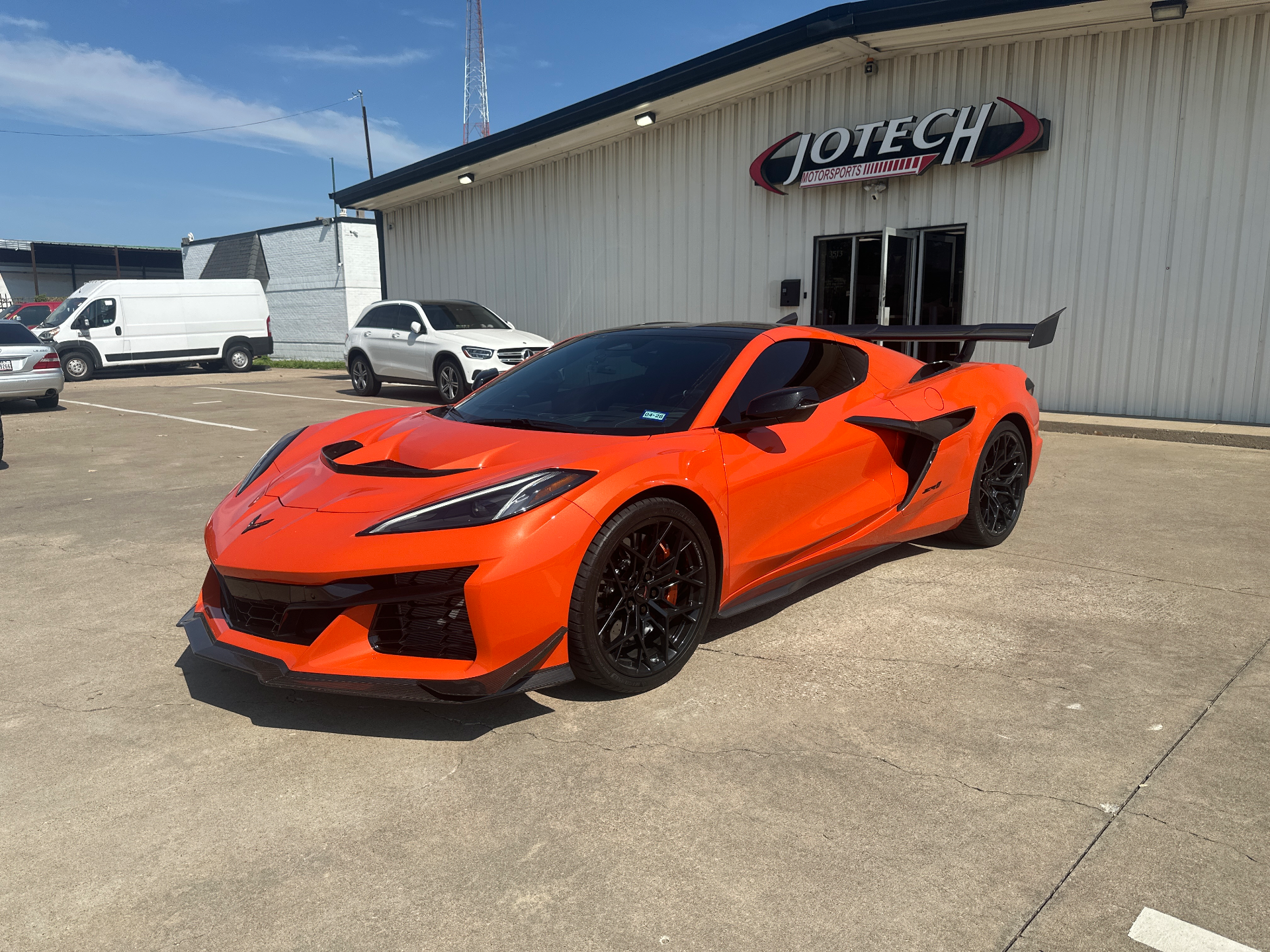 An orange Chevrolet Corvette C8 Z06 with a large rear wing and black wheels parked in front of the Jotech Motorsports building.