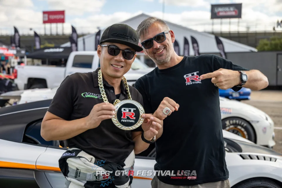Two men smiling and holding a GT-R Festival trophy at an outdoor car event, with cars in the background.