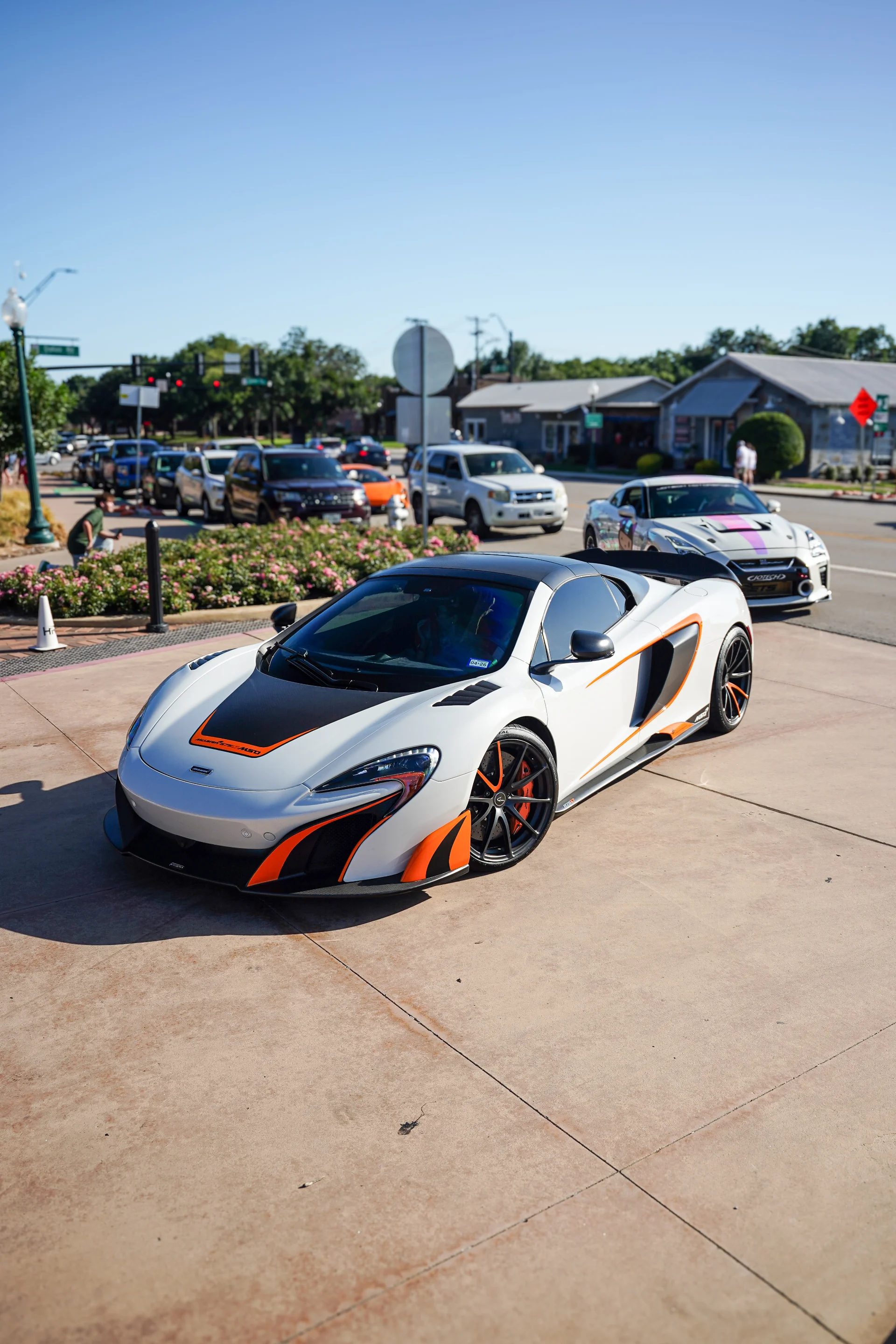 A white McLaren supercar with orange accents parked on a paved area at a car meet, with other vehicles and buildings in the background.