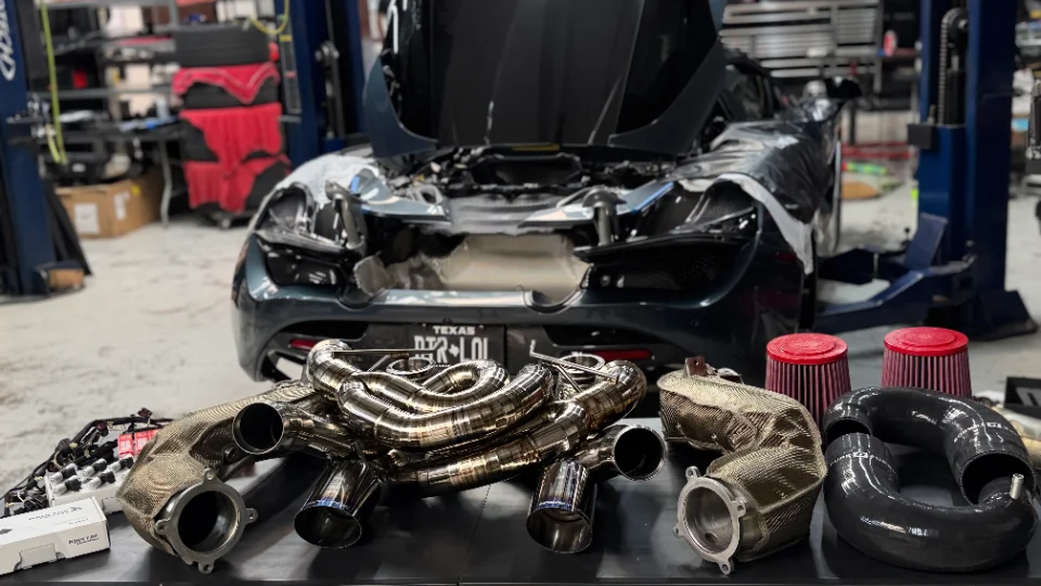 A McLaren supercar undergoing performance upgrades in a shop, with aftermarket exhaust headers, air filters, and boost pipes laid out.
