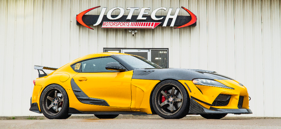 A modified yellow and black Toyota Supra with carbon fiber aero and aftermarket wheels parked in front of Jotech Motorsports.