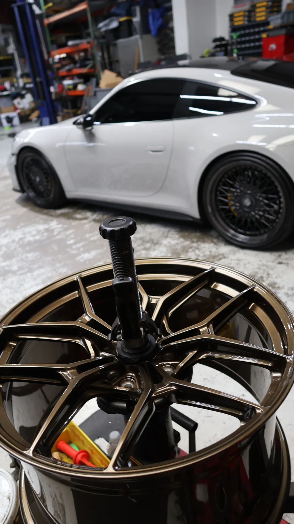 A bronze aftermarket wheel on a stand in a workshop, with a white Porsche 911 visible in the blurred background.