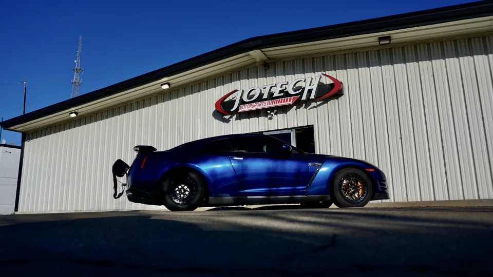 A blue Nissan GT-R with drag wheels and a parachute mount parked outside the Jotech Motorsports facility.