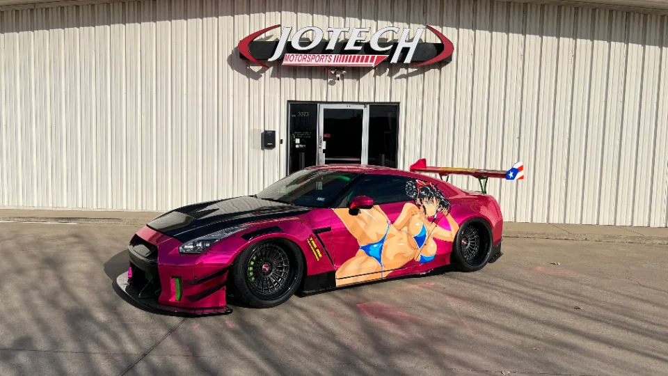 A vibrant pink Nissan GT-R with an anime-style itasha wrap and a large rear wing, parked in front of the Jotech Motorsports facility.