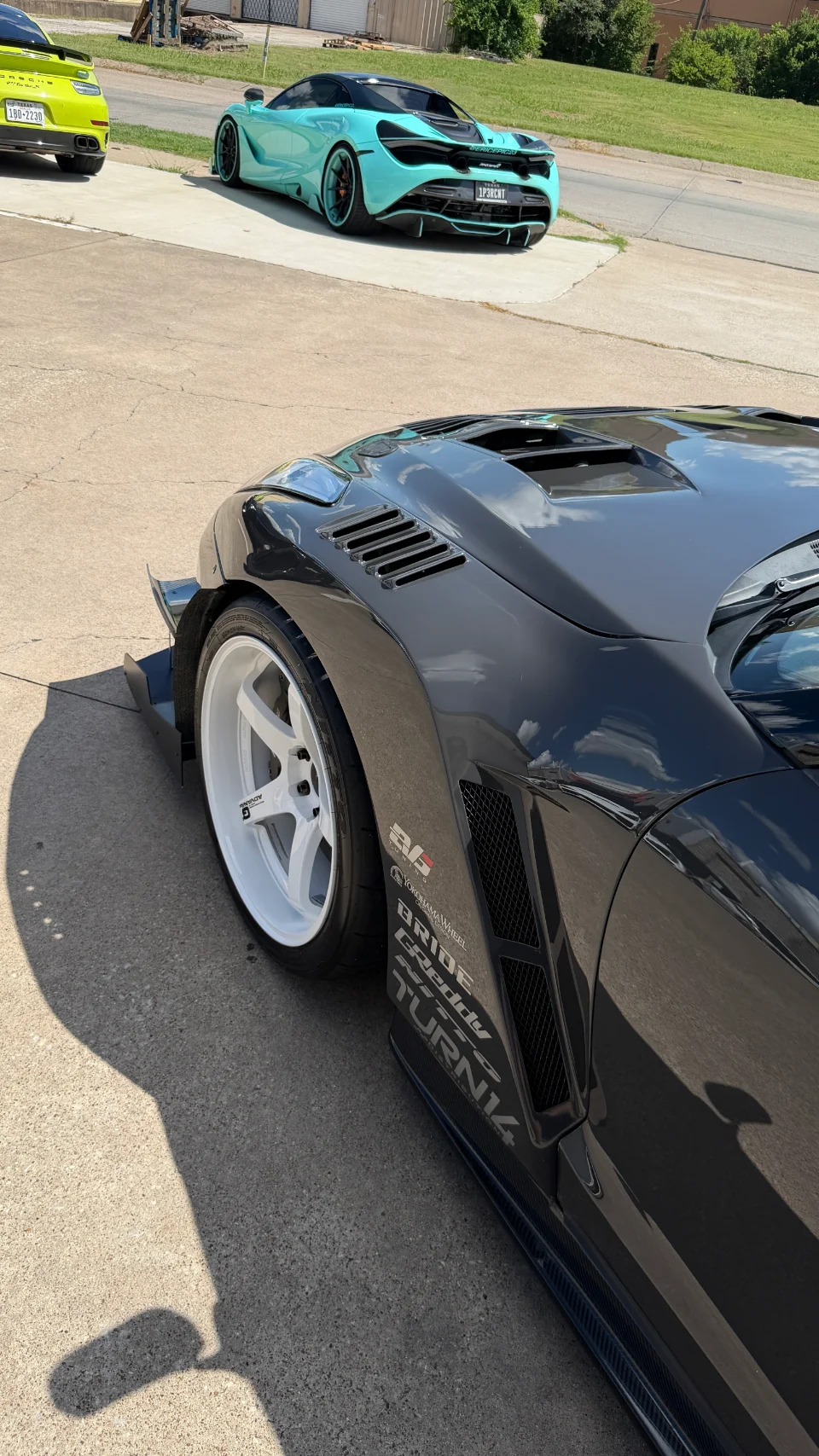 A black widebody car with white Advan wheels and custom fender vents parked on a street with a teal McLaren 720S and yellow Porsche 911 in the background.