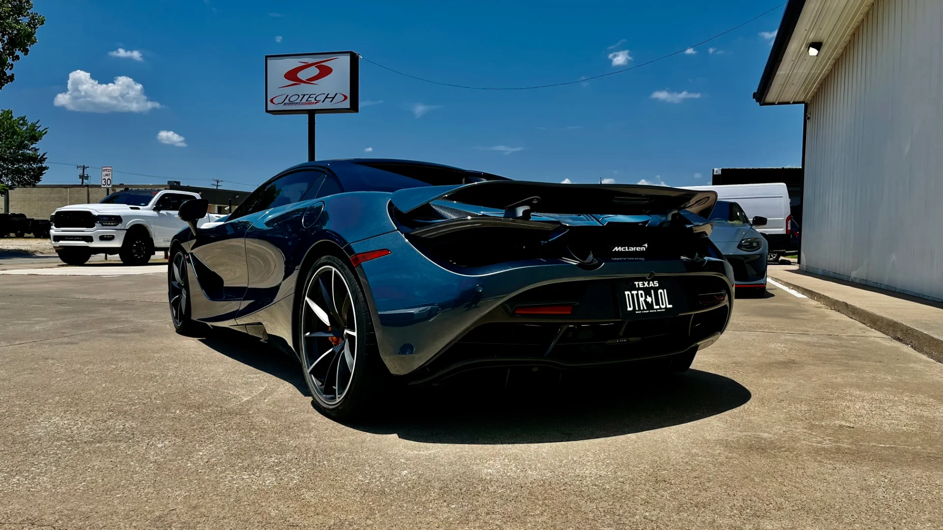 A blue McLaren 720S parked outside Jotech Motorsports shop under a clear blue sky, with a white truck in the background.