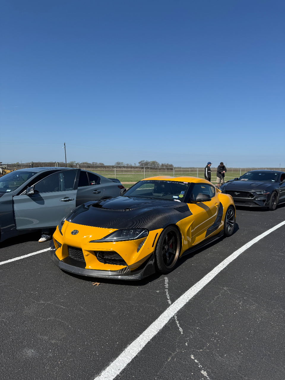 A highly modified yellow Toyota Supra with extensive carbon fiber bodywork, including a hood and widebody fenders, parked at a track event.