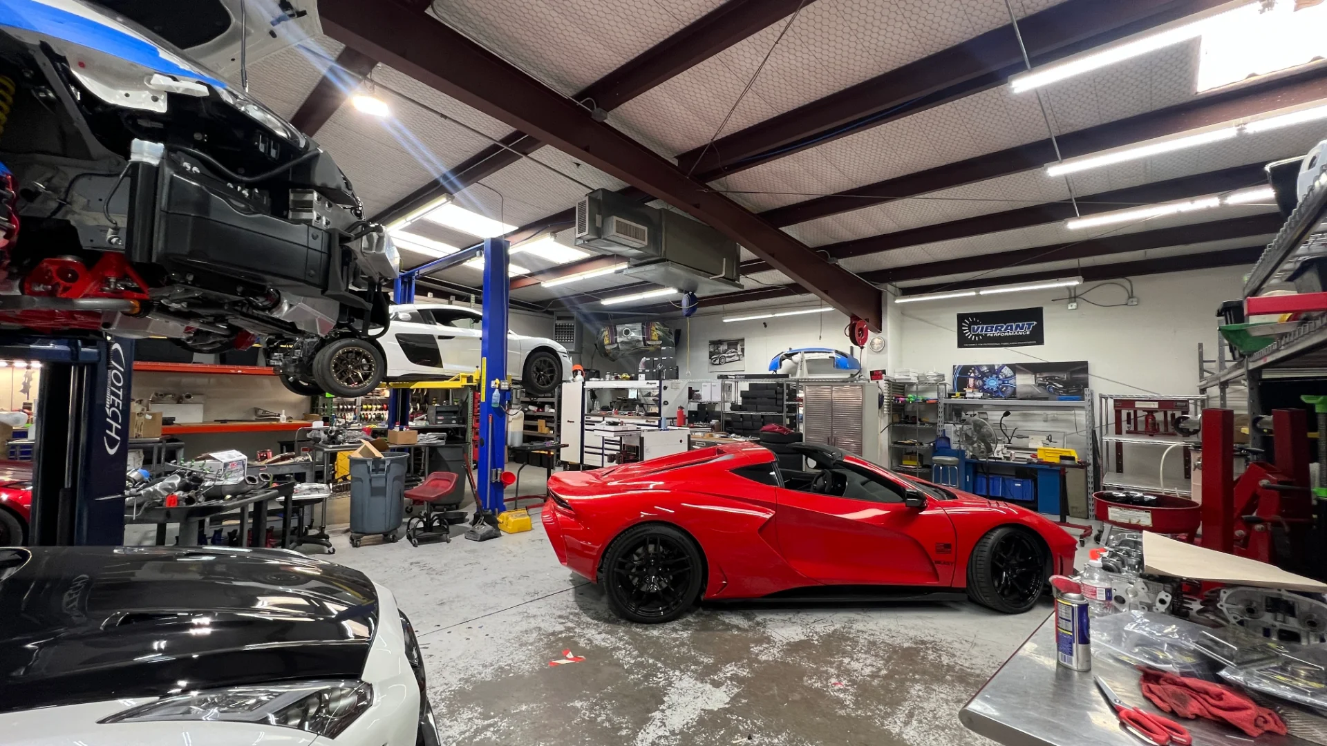 Interior view of a Jotech Motorsports performance shop with multiple cars on lifts and a red Corvette C8 parked on the floor.