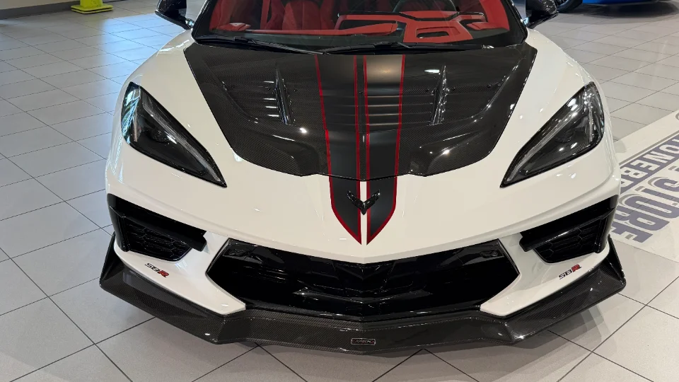 A white C8 Corvette with a carbon fiber hood and red racing stripes, featuring a prominent front splitter, displayed indoors.