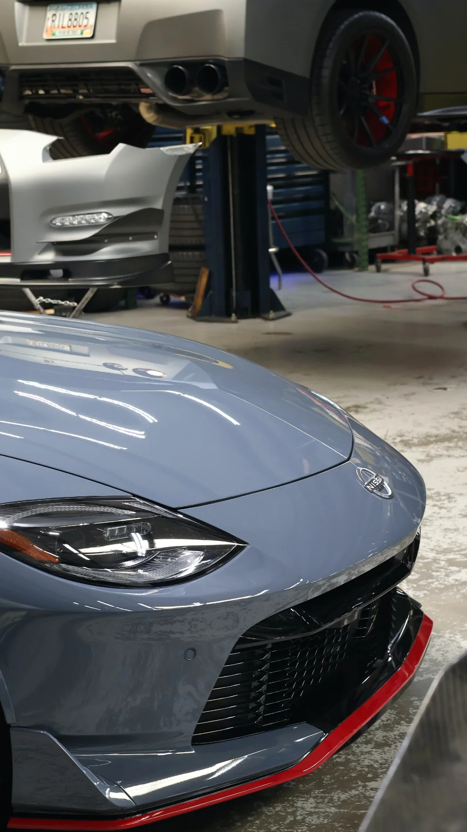 A grey Nissan Z with a red front splitter in the foreground, and a grey Nissan GT-R on a lift in a performance tuning shop.