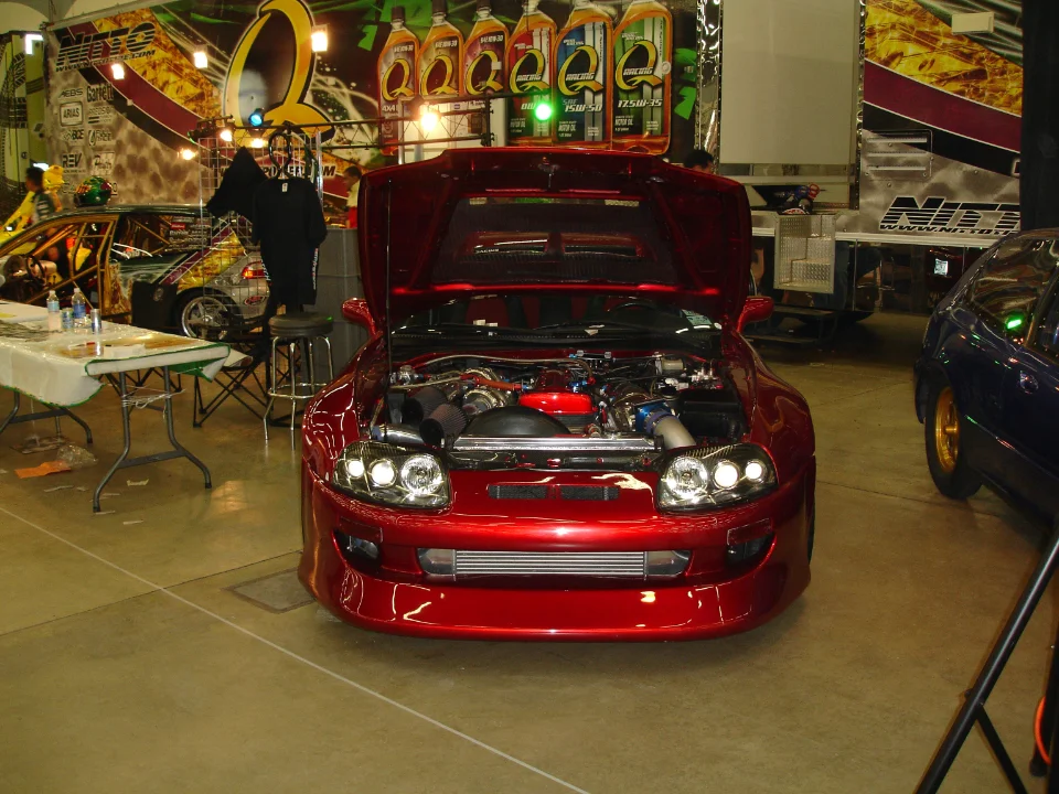 A red modified Toyota Supra with its hood open, showcasing a large turbocharger and intercooler setup at an indoor car show.
