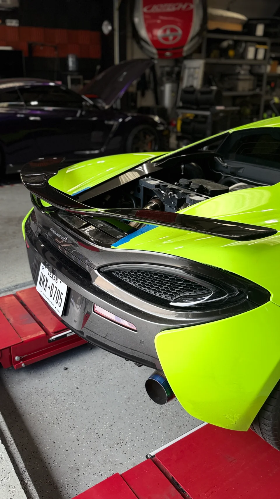 Rear view of a neon green McLaren 570S in a shop with its engine bay open, showcasing an aftermarket exhaust and rear wing.