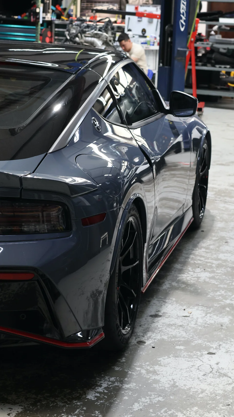 A grey Nissan Z sports car with black wheels parked in a Jotech Motorsports shop with a technician working in the background.