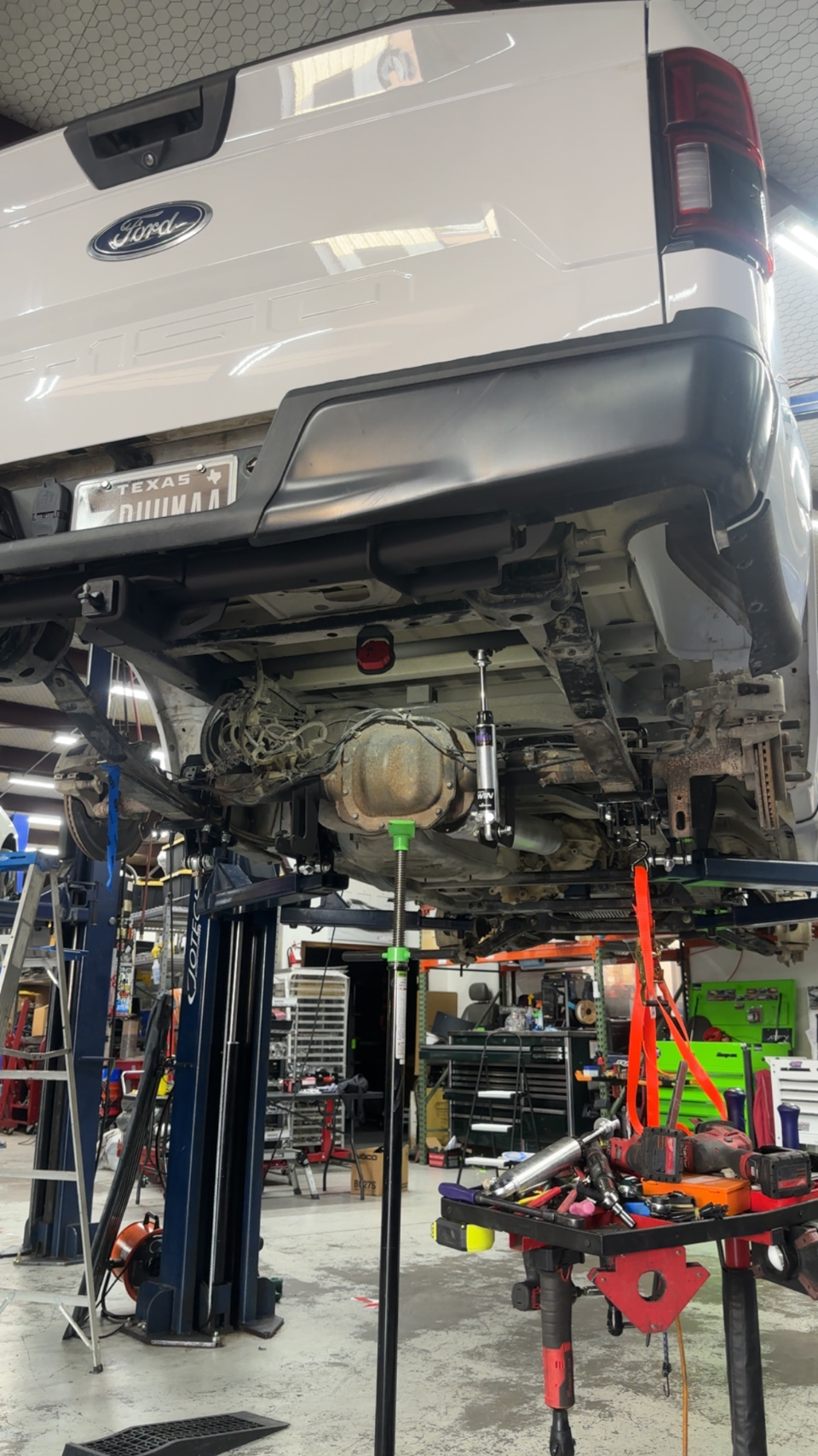 A white Ford truck on a lift in a shop, with the rear suspension and differential visible during an upgrade or repair.