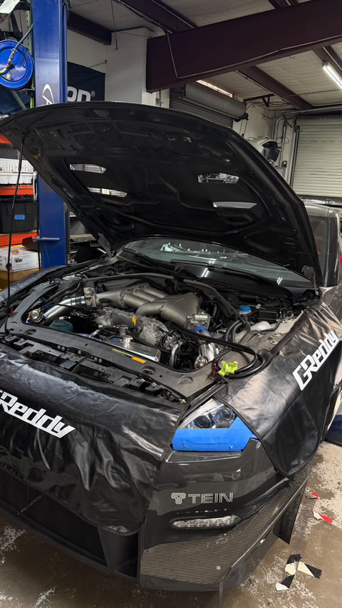 Nissan GT-R with its hood open in a Jotech Motorsports shop, showing the engine bay and GReddy fender covers.