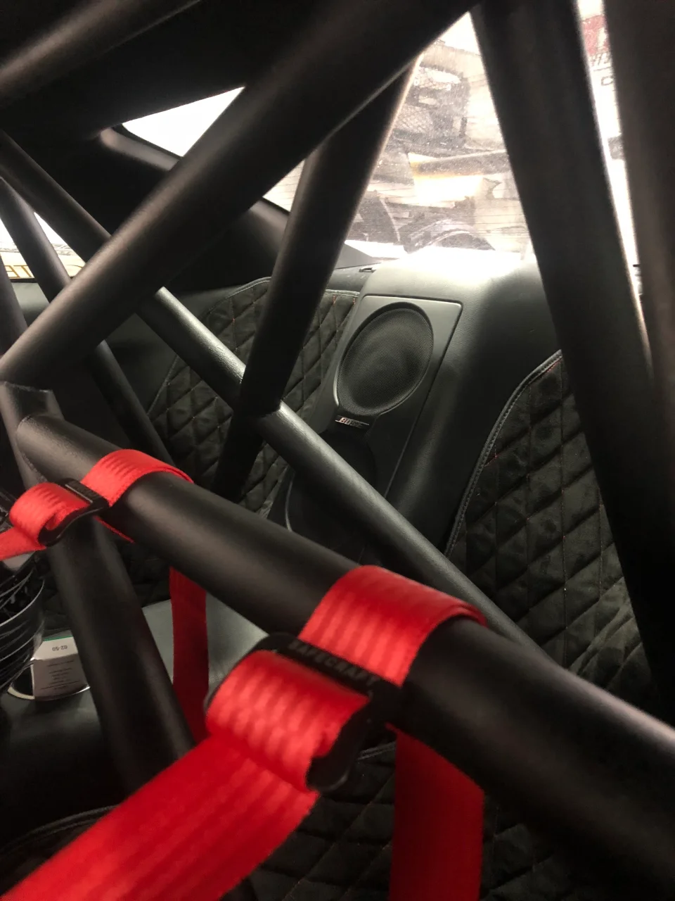 Interior view of a car showing a black roll cage, red racing harnesses, and quilted door panel with a Bose speaker.