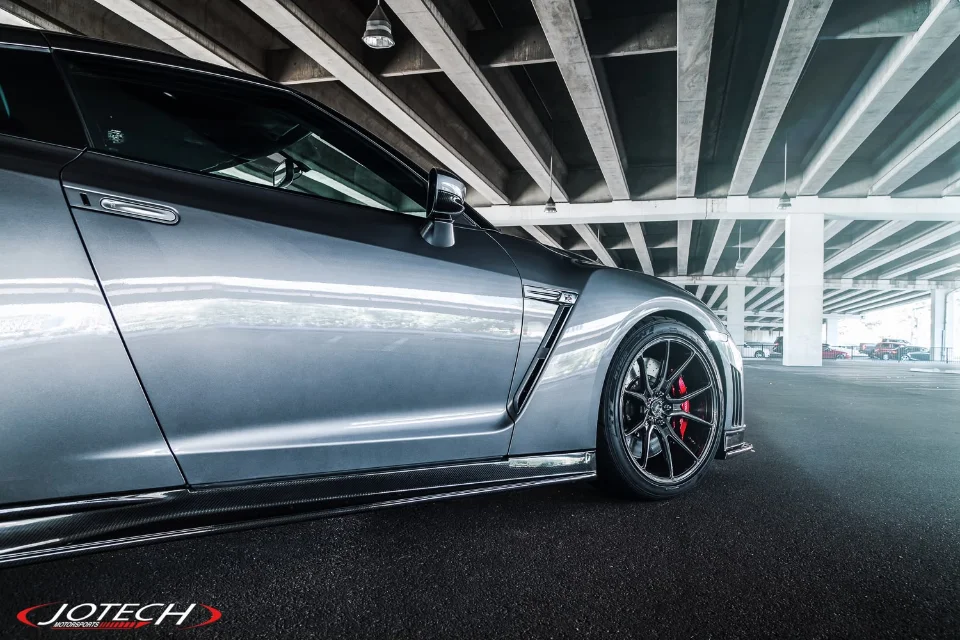 Grey Nissan GT-R with aftermarket black wheels and red brake calipers, featuring carbon fiber side skirts, parked under an overpass.