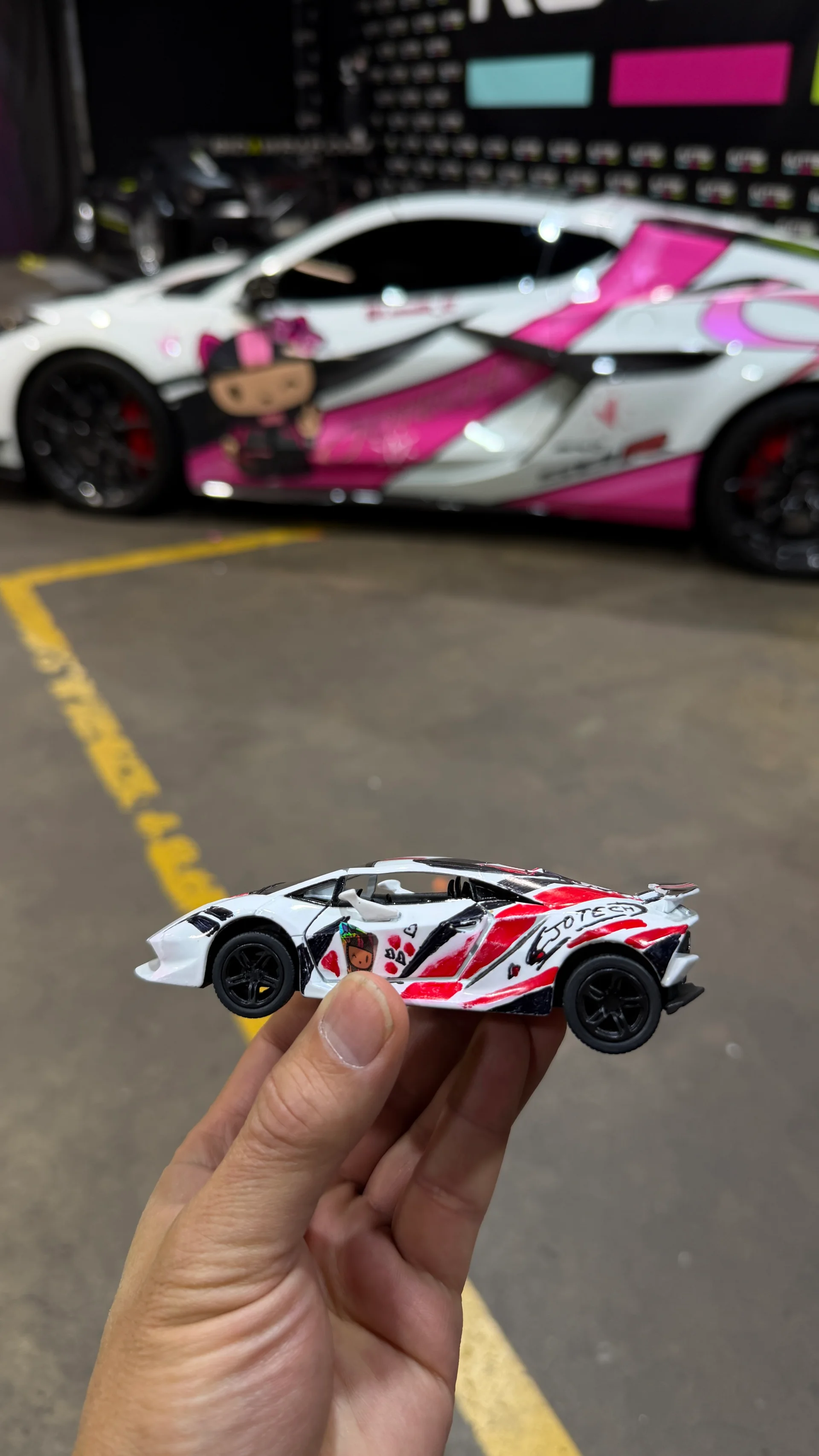A hand holds a miniature Lamborghini Huracan toy car with a pink and white livery, matching a real Lamborghini Huracan parked out of focus in the background at 