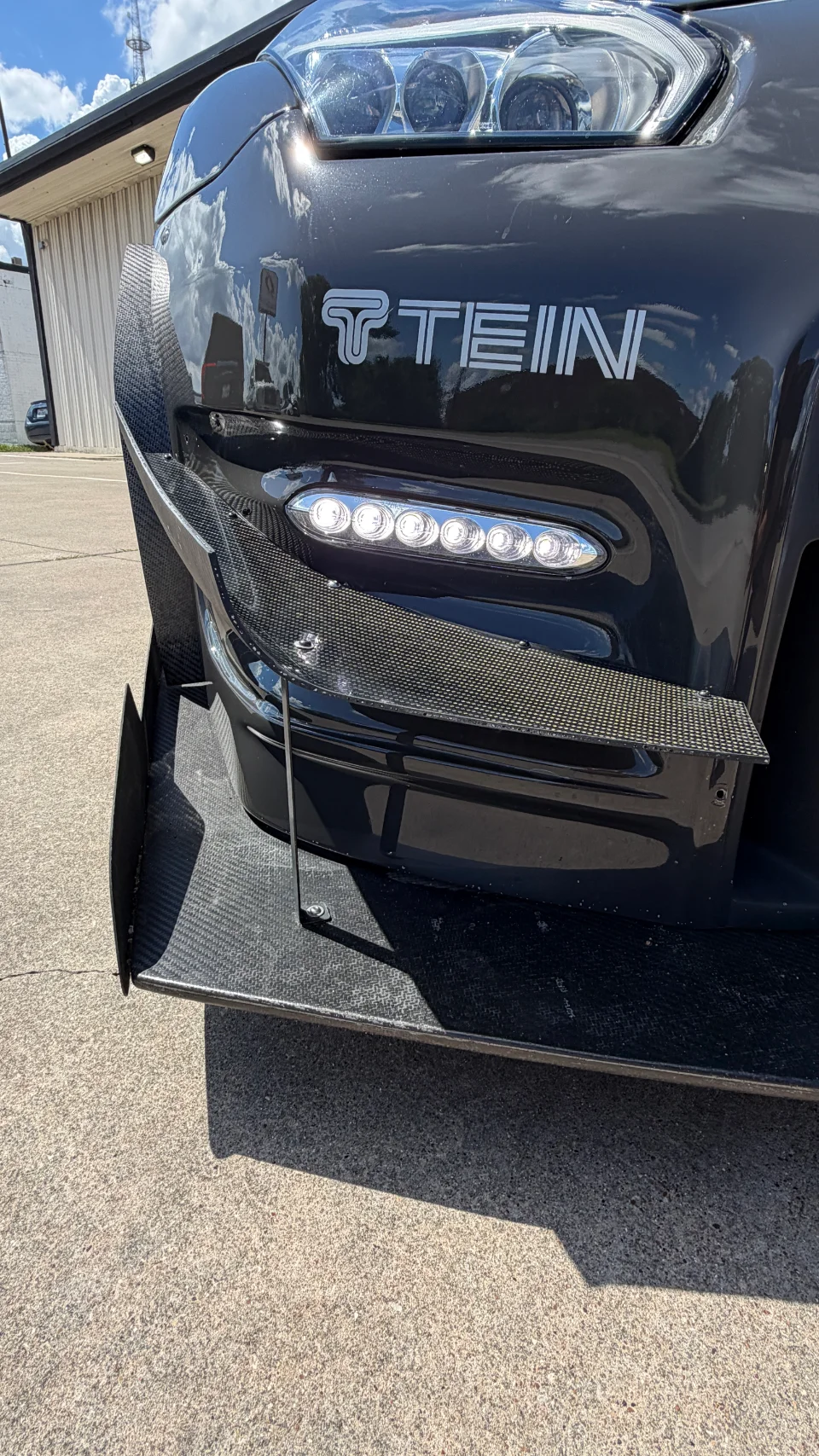 A close-up of the front bumper of a black car featuring a carbon fiber front splitter, canards, and LED daytime running lights.