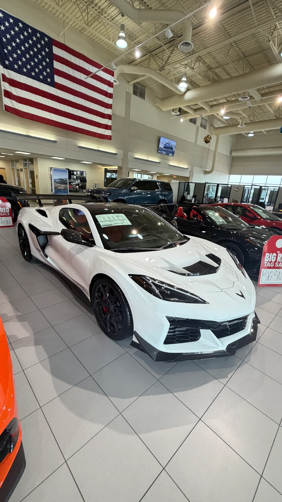 A white Chevrolet Corvette C8 Z06 sports car displayed in a bright dealership showroom with an American flag.