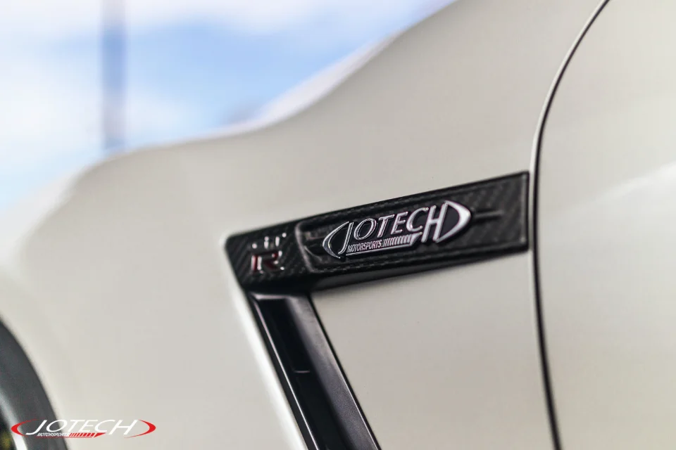 A close-up of a white Nissan GT-R fender with a Jotech Motorsports carbon fiber badge and vent.